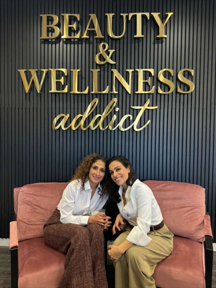 Dr. Sheila Omrani and Nurse Lisa Farazmand seated together beneath the gold Beauty and Wellness addict signage at their Great Neck practice.